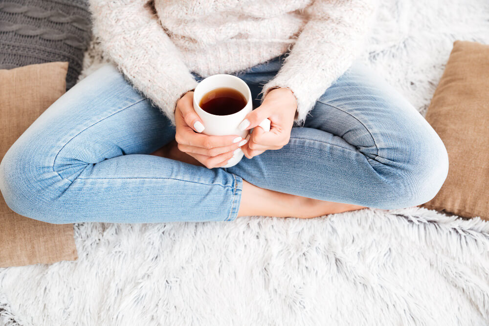 cropped image woman sweater jeans holding tea cup while sitting carpet indoors 1